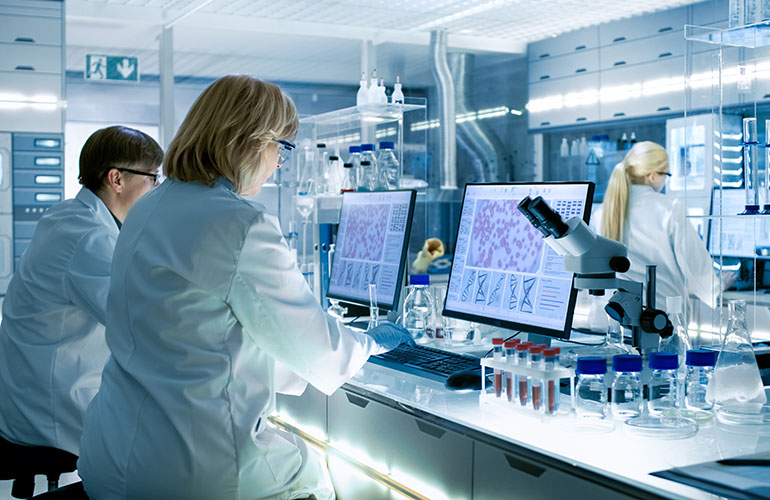 Female and Male Scientists Working on their Computers In Big Modern Laboratory. Various Shelves with Beakers, Chemicals and Different Technical Equipment is Visible.