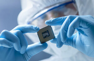 In Ultra Modern Electronic Manufacturing Factory Design Engineer in Sterile Coverall Holds Microchip with Gloves and Examines it.