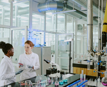 Scientists in white lab coats discussing experimental results in modern, well-equipped laboratory surrounded by various scientific instruments and glassware
