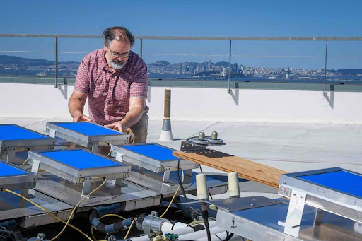 Caption: Howdy Goudey checks the levels on an array testing apparatus measuring a type of Passive Radiative Cooling Film on the roof of Building 59 at Berkeley Lab. Credit: Thor Swift/Berkeley Lab