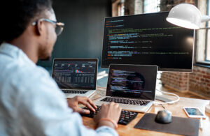 Young african male programmer writing program code sitting at the workplace with three monitors in the office. Image focused on the screen
