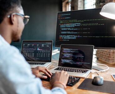 Young african male programmer writing program code sitting at the workplace with three monitors in the office. Image focused on the screen