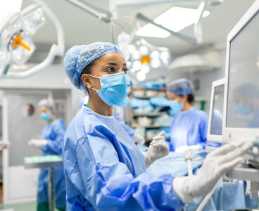 Anesthetist Working In Operating Theatre Wearing Protecive Gear checking monitors while sedating patient before surgical procedure in hospital