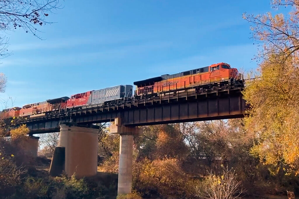A BNSF freight consist led by diesel locomotive #3965 and featuring Wabtec’s FLXdrive 100% battery-electric locomotive (#3000) situated between conventional Tier 4 diesel units crosses a bridge in California during revenue service pilot testing.