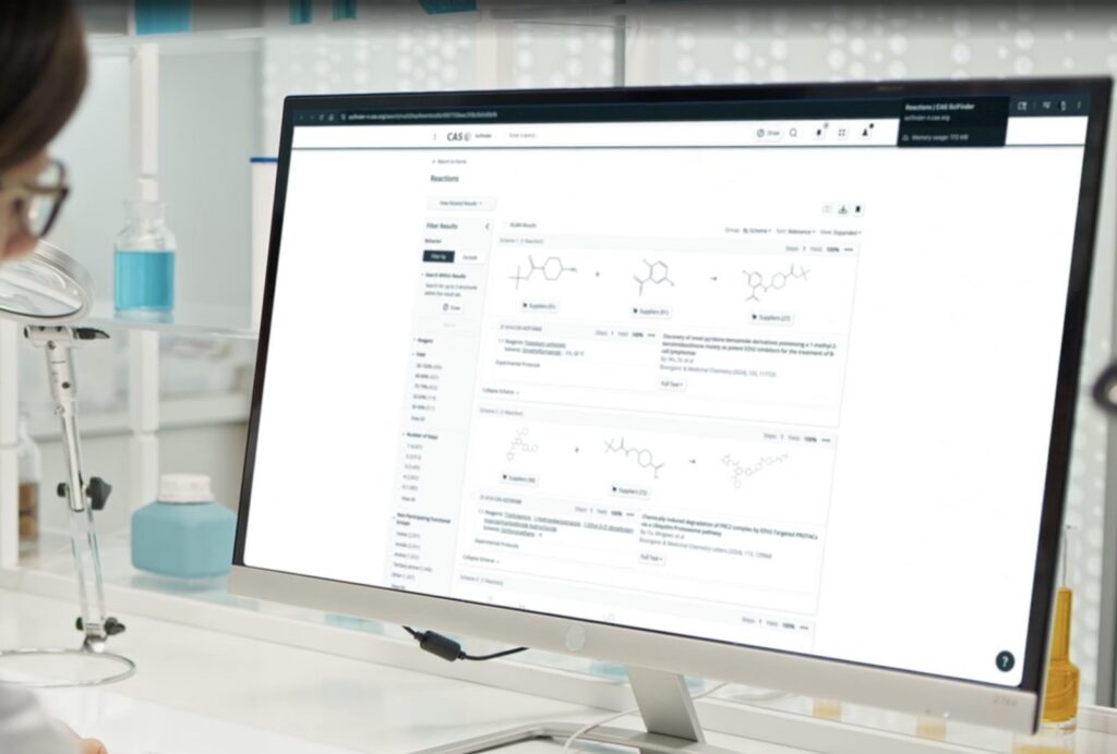 A scientist in a lab looks at CAS SciFinder reaction search results on a desktop monitor.