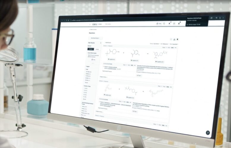 A scientist in a lab looks at CAS SciFinder reaction search results on a desktop monitor.